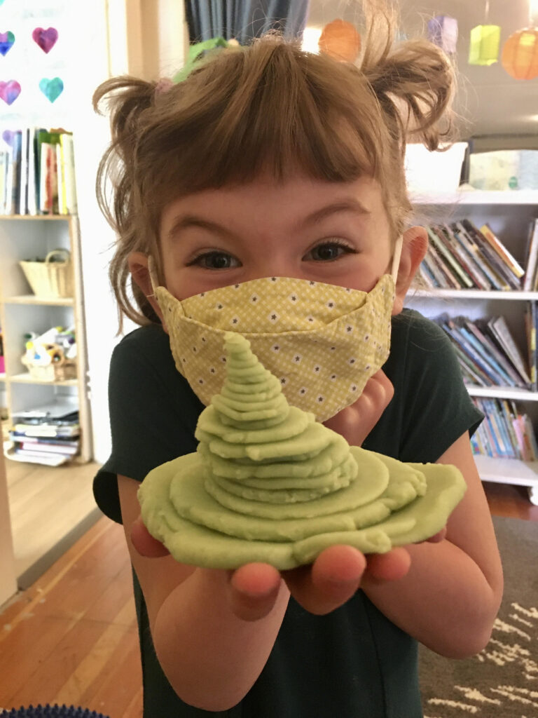 child with a mask holding a stack of play dough for the camera and smiling